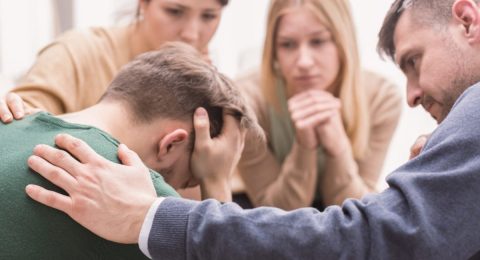 Close-up of a devastated young man holding his head in his hands and friends supporting him during group therapy