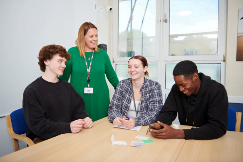 myth busting: three young people sat around a table discussing and laughing. One member of staff in a green dress stood behind them smiling