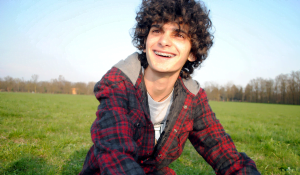 Dylan with curley brown hair and braces smiling at the camera, sat in a grass field