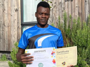 Nader stood in the garden in a blue sports tshirt holding up two certificates of achievement