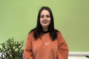 Georgia, smiling stood against a green background with a plant to her side