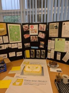 a white and yellow celebration cake in front of a display about the YMCA downsLink group What is Sexual Exploitation (WiSE) project