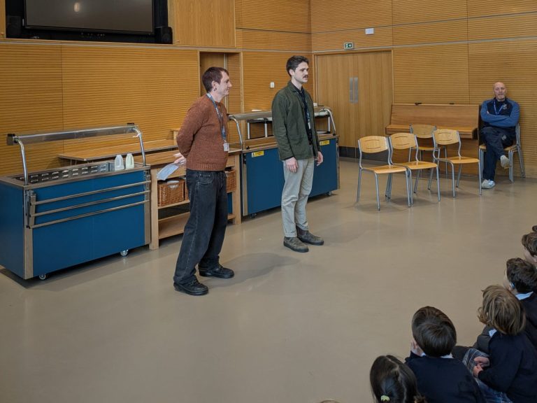 two men standing in front of an assembly of seated children mostly off screen