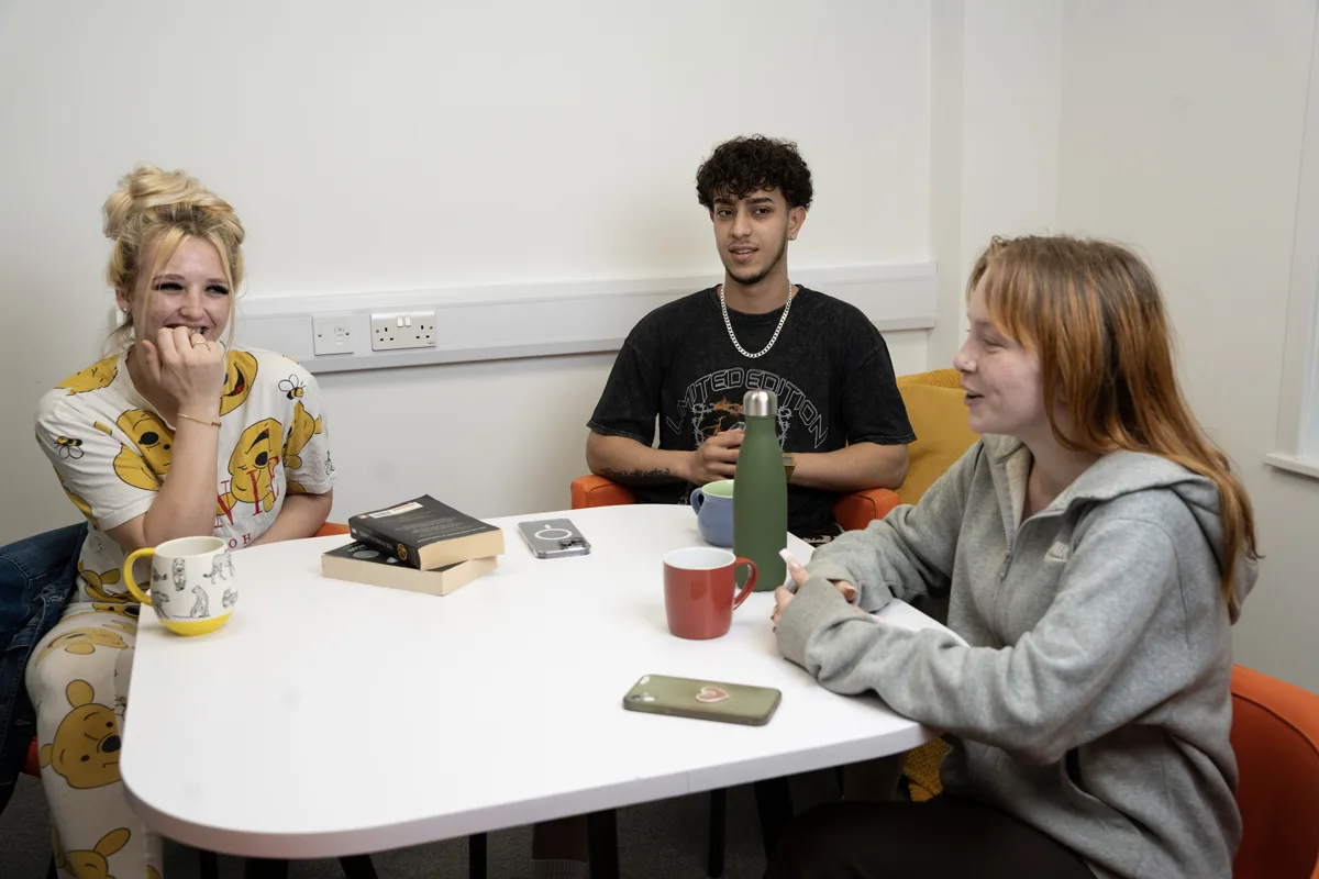 3 young people, 2 female one male, sat around a table with drinks and books