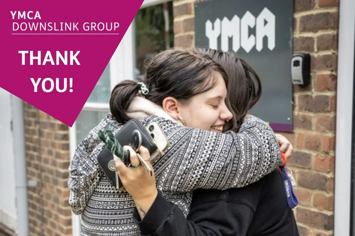 two women embracing in front of a YMCA sign. The one with a visible face is smiling.