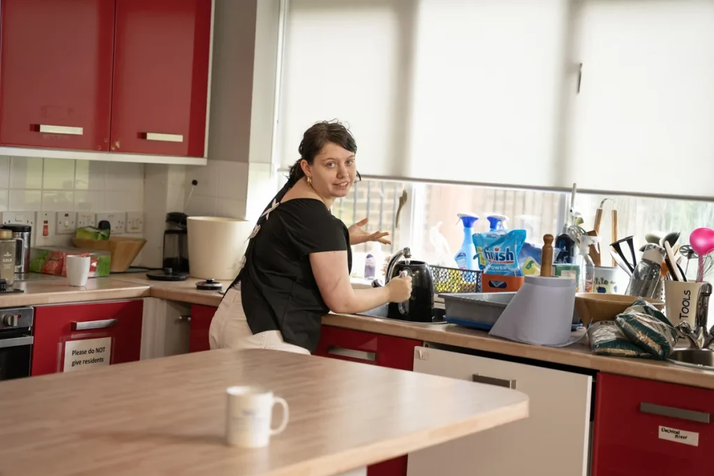 young woman in kitchen, filling a kettle, looking over her shoulder at camera