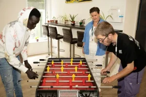 2 young men playing table football, watched by an adult wearing a lanyard