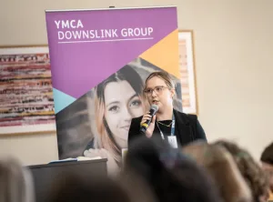 woman with glasses and microphone in front of a YMCA DownsLink group branded banner