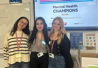 3 young women stood in front of a screen reading 'Mental Health CHAMPIONS'