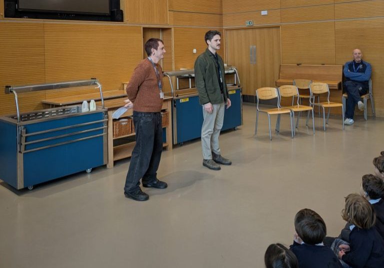 two men standing in front of an assembly of seated children mostly off screen