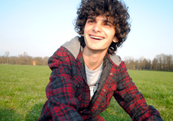 Dylan with curley brown hair and braces smiling at the camera, sat in a grass field