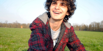 Untitled design (13) Dylan with curley brown hair and braces smiling at the camera, sat in a grass field