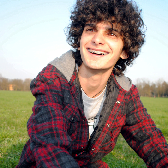 Untitled design (13) Dylan with curley brown hair and braces smiling at the camera, sat in a grass field