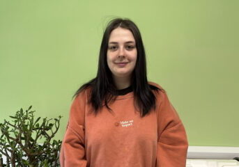 Georgia, smiling stood against a green background with a plant to her side