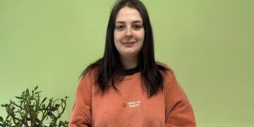 Worthing Foyer_Resident Georgia_March 2024 for cover image Georgia, smiling stood against a green background with a plant to her side
