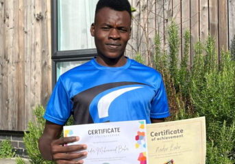 Nader stood in the garden in a blue sports tshirt holding up two certificates of achievement