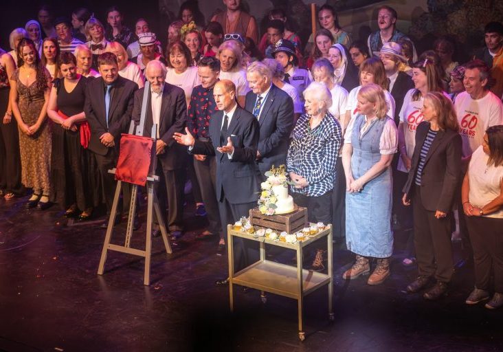 a crowd of people listening to a man stood at their centre near a tiered celebration cake